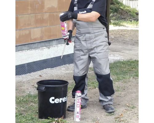 Remeselník v pracovnom odeve drží aplikačnú pištoľ s lepidlom Ceresit CT 84 nad vedrom vonku.
