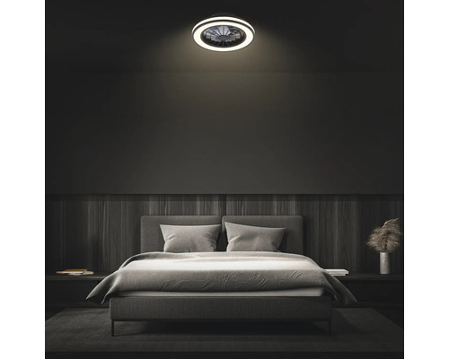 Moderný stropný ventilátor s LED osvetlením v tmavo zariadenej spálni nad sivou čalúnenou posteľou.
