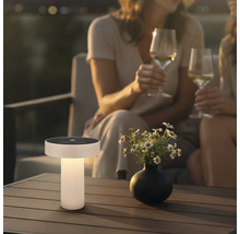 Moderná solárna stolná lampa na drevenom terasovom stole v záhrade za súmraku s kvetmi a ľuďmi v pozadí.