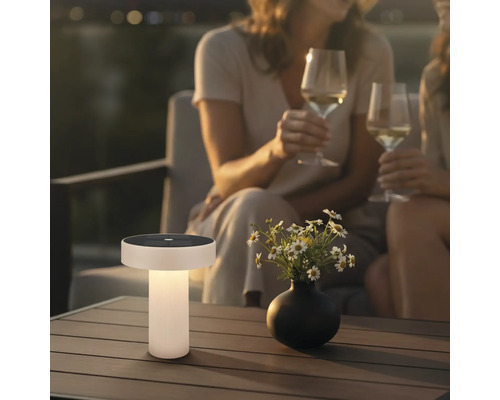 Moderná solárna stolná lampa na drevenom terasovom stole v záhrade za súmraku s kvetmi a ľuďmi v pozadí.