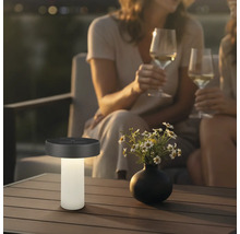 Moderná solárna stolná lampa na terasovom stole vedľa vázy s kvetmi, v pozadí dve osoby s pohármi vína vo večernej atmosfére.