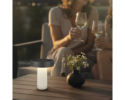 Moderná solárna stolná lampa na terasovom stole vedľa vázy s kvetmi, v pozadí dve osoby s pohármi vína vo večernej atmosfére.