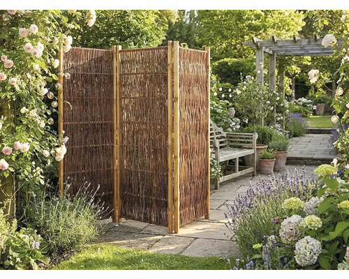 Trojdielna skladacia zástena z prútia v záhrade s kamennou dlažbou, kvetmi a drevenou lavičkou.