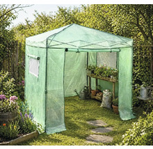 Priechodný fóliovník v záhrade s dreveným regálom, rastlinami v kvetináčoch, kanvicou a vreciom so zeminou na trávniku.