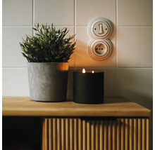 Vypínač a zásuvka v retro štýle na kachľovej stene vedľa rastliny a sviečky na drevenej komode.