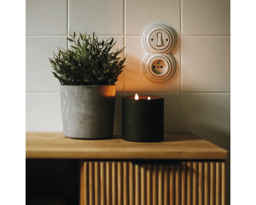 Vypínač a zásuvka v retro štýle na kachľovej stene vedľa rastliny a sviečky na drevenej komode.