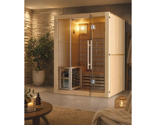 Interiérová sauna z dreva s preskleným čelom a dverami vo svetlej wellness miestnosti s kamennou stenou, izbovou rastlinou a príslušenstvom.