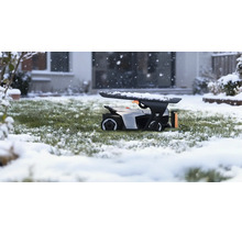 Robotická kosačka v garáži na zasneženom trávniku počas sneženia pred domom.
