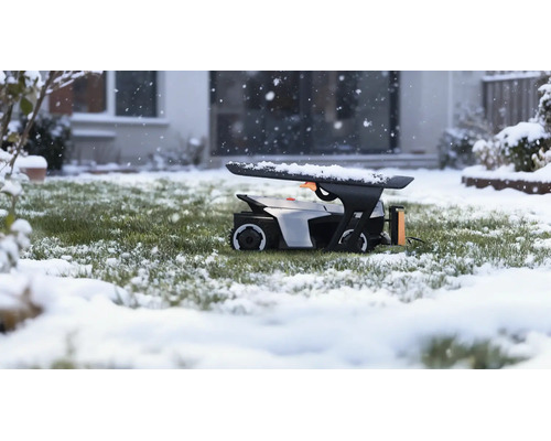 Robotická kosačka v garáži na zasneženom trávniku počas sneženia pred domom.