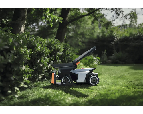 Robotická kosačka s príslušenstvom na trávniku v záhrade vedľa živého plota.