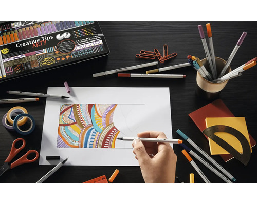 Ruka kreslí farebné vzory fixkou Stabilo Creative Tips na papier, obklopená rôznymi kresliacimi potrebami a kovovým puzdrom.