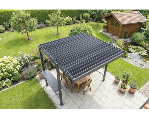 Voľne stojaca pergola s lamelovou strechou na svetlej terase v záhrade, vrátane dreveného nábytku, priľahlého jazierka a záhradného domčeka v pozadí.
