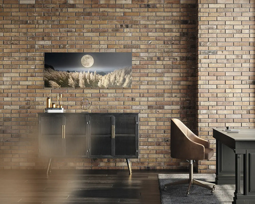 Panoramatický nástenný obraz s úplnkom nad okrasnými trávami na tehlovej stene nad čiernou komodou v modernej izbe s hnedou kancelárskou stoličkou.