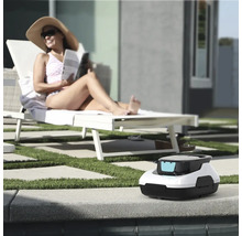 Bazénový robot na okraji bazéna, v pozadí žena na ležadle vo svetlej záhrade s trávnikom a dlažbou.