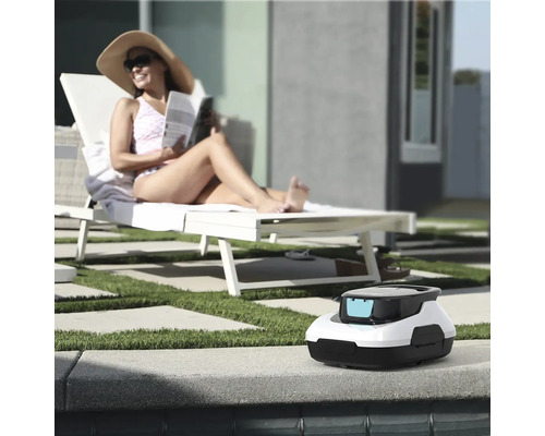 Bazénový robot na okraji bazéna, v pozadí žena na ležadle vo svetlej záhrade s trávnikom a dlažbou.