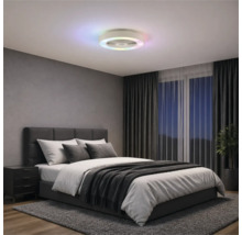 Moderný stropný ventilátor s farebným LED kruhom v spálni so sivou čalúnenou posteľou, tmavými závesmi a kobercom.
