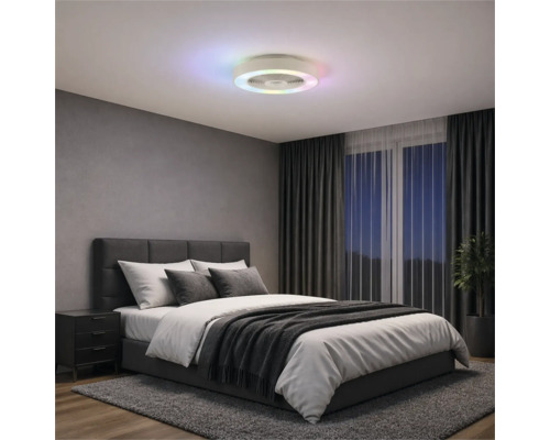 Moderný stropný ventilátor s farebným LED kruhom v spálni so sivou čalúnenou posteľou, tmavými závesmi a kobercom.