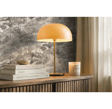 Moderná stolná lampa s okrúhlym tienidlom na drevenej komode pred lesnou tapetou, doplnená knihami a vonnou sviečkou. Svetlé a usporiadané.
