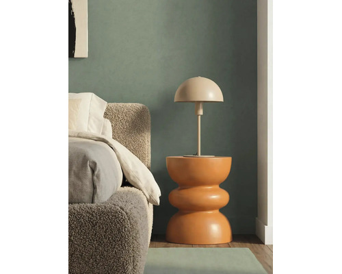 Stolná lampa v tvare huby na sochárskom odkladacom stolíku vedľa postele s čelom z buklé tkaniny pred tmavozelenou stenou.