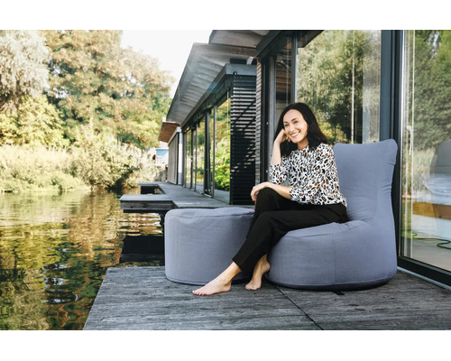 Žena sedí na sivom outdoorovom sedacom vaku s ladiacou podnožkou na drevenej terase pri vode pred moderným domom.