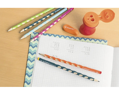 Písací stôl s ergonomickými ceruzkami Stabilo EASYgraph, gumou, orezávačom so zásobníkom a zošitom s matematickými výpočtami.