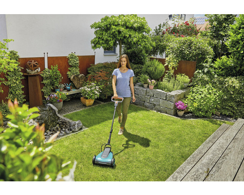 Žena používa kosačku na trávu v udržiavanej záhrade so skalkou a bujnou výsadbou.