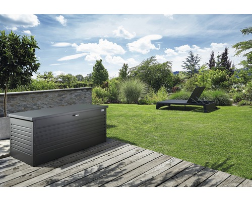 Záhradná scéna s kovovým úložným boxom na drevenej terase a lehátkom na trávniku