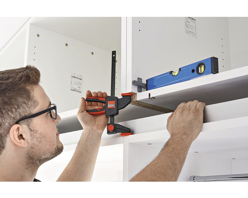 Osoba pripevňuje policu svorkou do bielej skrine.