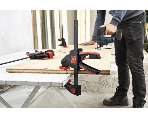 Obrábaný kus upevnený skrutkovými svorkami, zatiaľ čo ho osoba opracováva priamočiarou pílou.