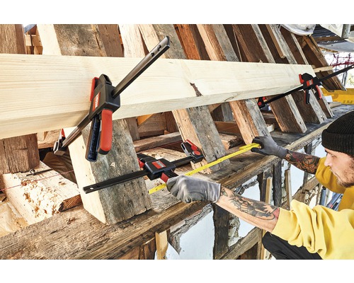 Remeselník meria kus dreva meracím pásmom, ktoré je upevnené skrutkovacími svorkami.