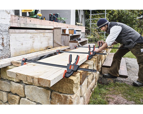 Remeselník upevňuje drevo k murovanej stene pomocou horizontálnej jednoručnej svorky.