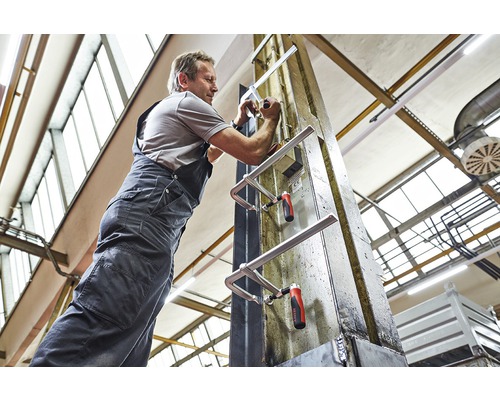 Muž upevňuje dve skrutkovacie svorky na kovový nosník v dielni.