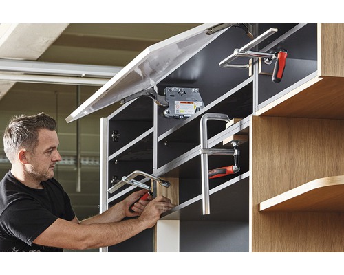 Muž pripevňuje čelo nábytku ku skrini pomocou svorky.