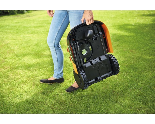 Osoba nesie robotickú kosačku cez trávnik.