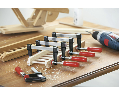 Drevený obrobok je upevnený skrutkovacími svorkami, vŕtačka v pozadí