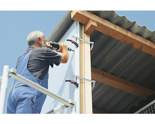 Remeselník pripevňuje fasádu k budove pomocou akumulátorového vŕtacieho skrutkovača, pričom ako podpora slúžia svorky.