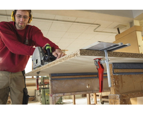 Remeselník reže drevenú dosku ponornou pílou, upevnenou skrutkovými svorkami na pracovný stôl