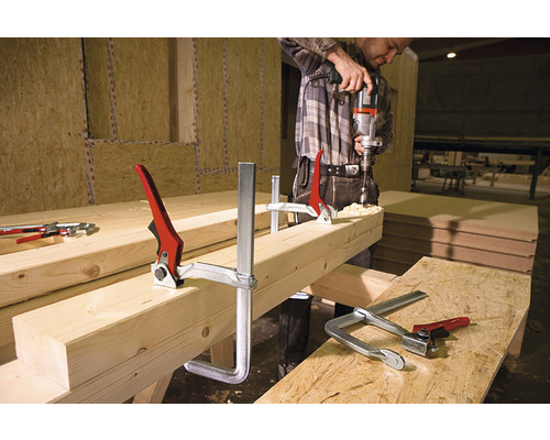 Muž vŕta vŕtačkou do kusu dreva, ktorý je upevnený skrutkovacími svorkami.