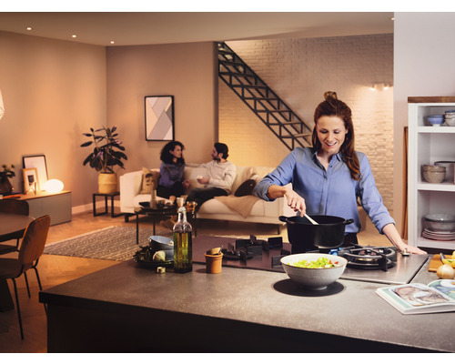 Svetlá otvorená obývacia kuchyňa so ženou, ktorá varí na sporáku a s ľuďmi sediacimi na pohovke v pozadí.
