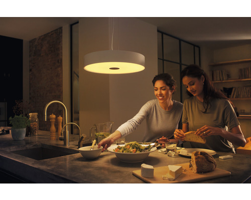 Dve ženy pripravujú jedlo v jasnej kuchyni pod okrúhlym závesným svetlom.