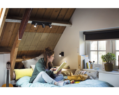 Útulná podkrovná izba s dreveným obkladom, vybavená stropným svietidlom s niekoľkými reflektormi a lampou na čítanie, kde na posteli sedí a číta mladá žena.