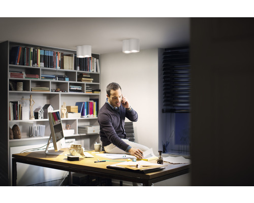 Muž sedí na stole v pracovni s knižnicou a stropnými svietidlami a telefonuje.