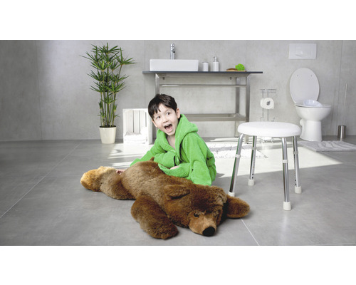 Svetlá kúpeľňa s chlapcom v župane sediacim vedľa medveďa na stoličke, s umývadlom, toaletou a rastlinou