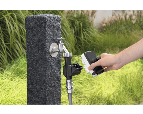 Záhradná scéna s vodovodným kohútikom na kamennom podstavci a osobou pripájajúcou zavlažovacie zariadenie