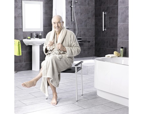 Muž v župane sedí na sprchovej stoličke v kúpeľni so sprchou, umývadlom a vaňou.
