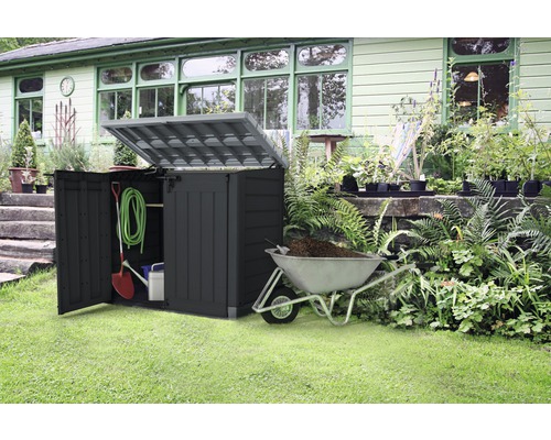 Záhradná scéna s otvoreným boxom na náradie, záhradným náradím a fúrikom naplneným zeminou.