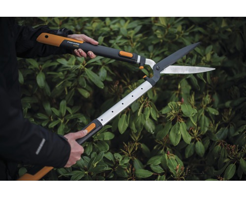 Osoba používa prevodové nožnice na živý plot pred zeleným živým plotom.