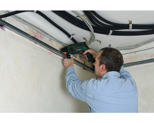 Muž vŕta príklepovou vŕtačkou do kovovej lišty na strope. Káble sú tiež viditeľné.