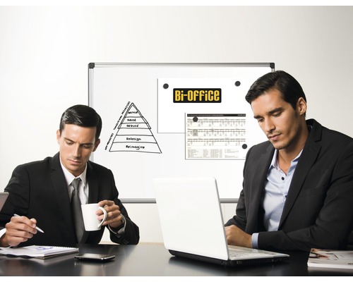 Dvaja muži sedia pri stole s notebookom pred prezentáciou na tabuli v kancelárii.