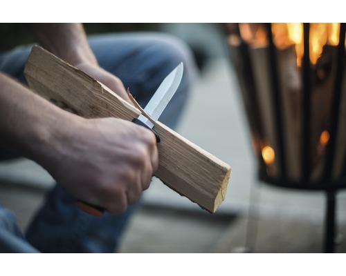 Osoba nožom orezáva drevo pred ohniskom.